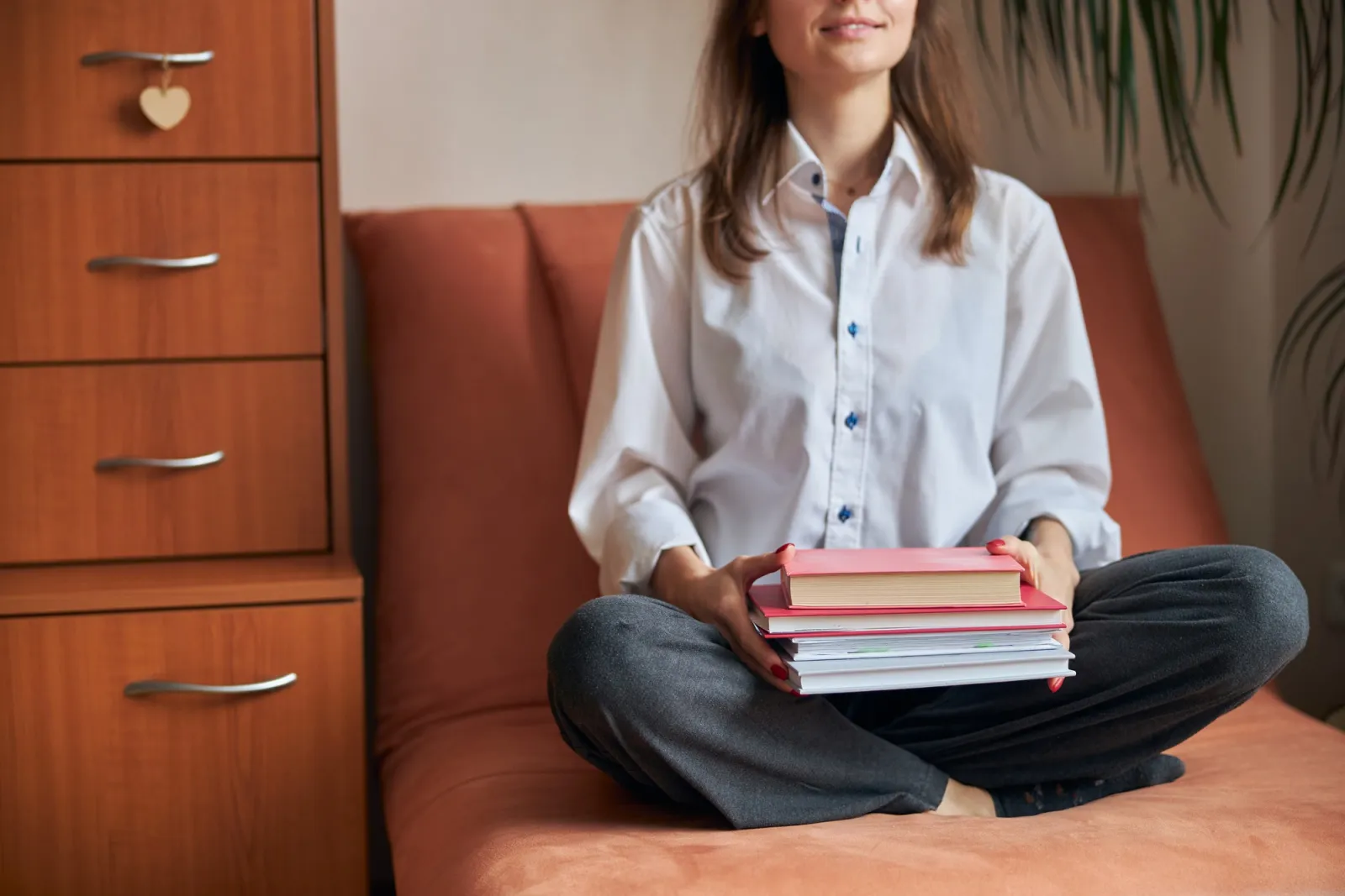 Frau sitzt im Schneidersitz auf einem Sofa und hält einen Stapel Bücher im Schoß